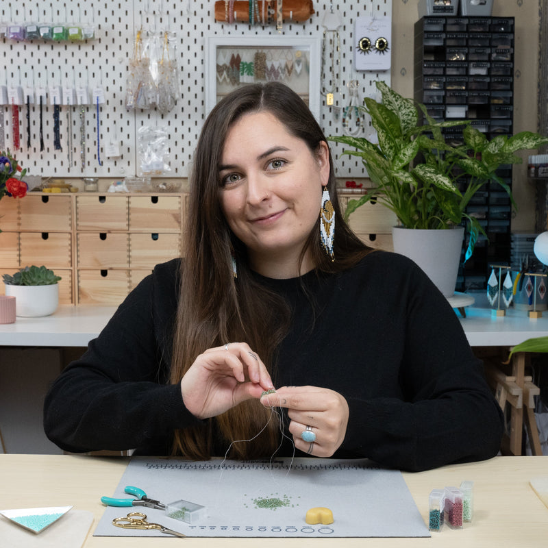 Woman sitting at a table in a craft or office setting with various items and plants around.